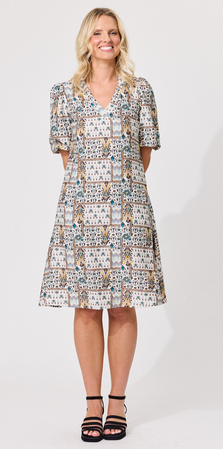 A woman modeling a knee-length patterned dress with short puff sleeves, a V-neckline, and a flared skirt, paired with black strappy heels against a plain light background.