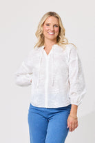 A smiling blonde woman wearing a white long-sleeve blouse with floral embroidery and drawstrings, paired with blue jeans, against a plain light gray studio background.