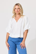 A blonde woman wearing a white short-sleeve blouse with a V-neck and puff sleeves, paired with blue jeans, standing against a light gray studio background.