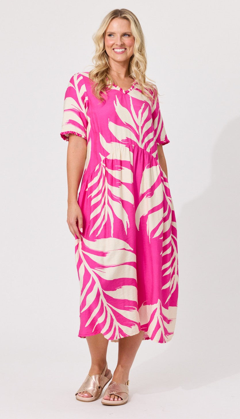 A woman modeling a short-sleeve midi dress with a vibrant pink and cream tropical leaf print, paired with metallic rose gold sandals, against a plain white studio background.