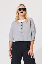 A woman modeling a cropped black and white gingham button-front jacket with three-quarter sleeves, paired with dark trousers and black sunglasses, against a plain light gray background.