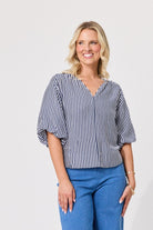 A woman modeling a navy and white vertically striped blouse with puff sleeves and a split V-neckline, paired with blue jeans, against a plain light gray background.