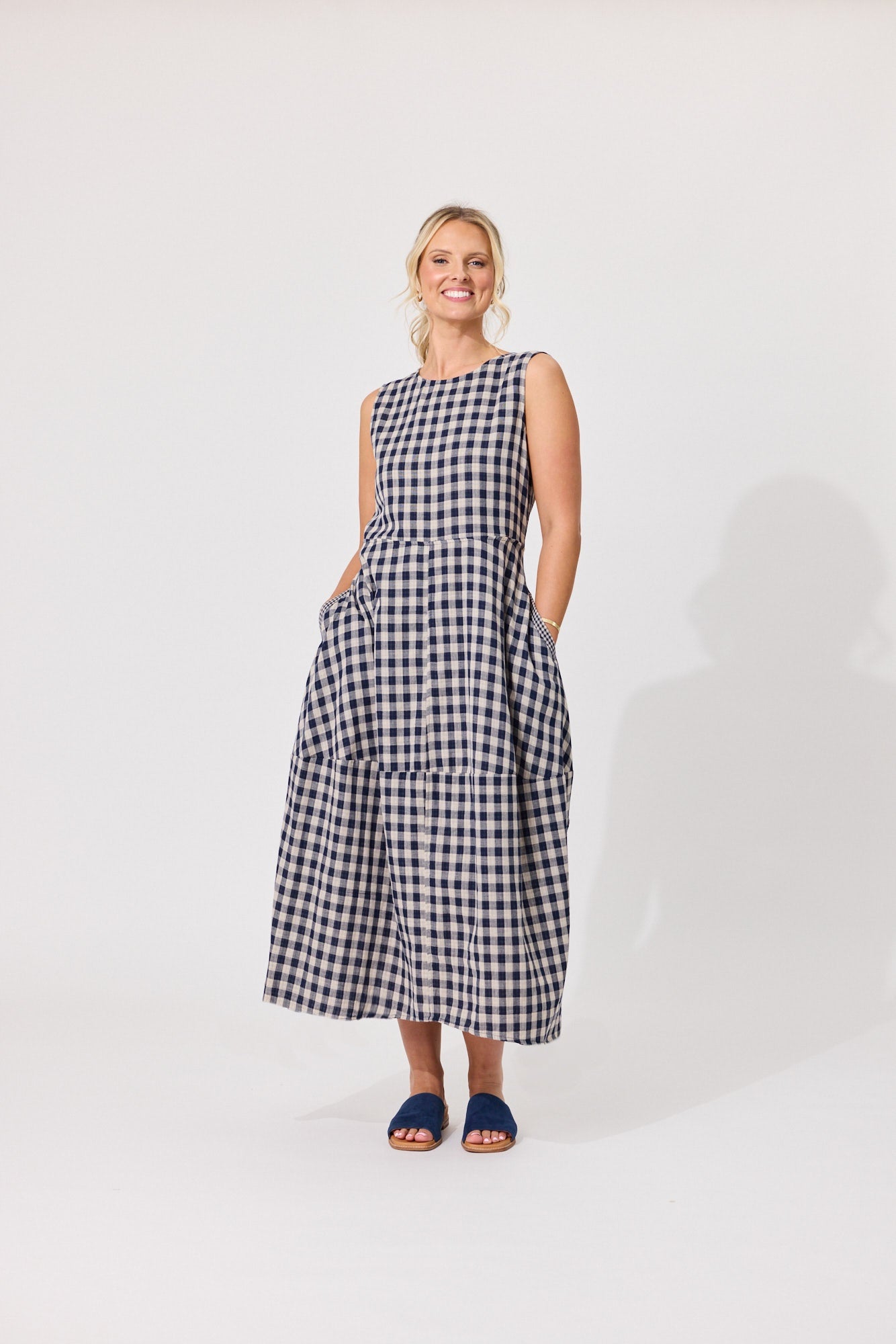 A woman modeling a sleeveless black and white gingham midi dress with side pockets, standing against a plain white studio background.