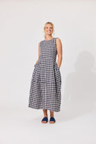 A woman modeling a sleeveless black and white gingham midi dress with side pockets, standing against a plain white studio background.