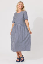 A woman modeling a navy and white vertically striped midi dress with short puff sleeves and a gathered waist, paired with dark blue slide sandals, against a plain light gray studio background.