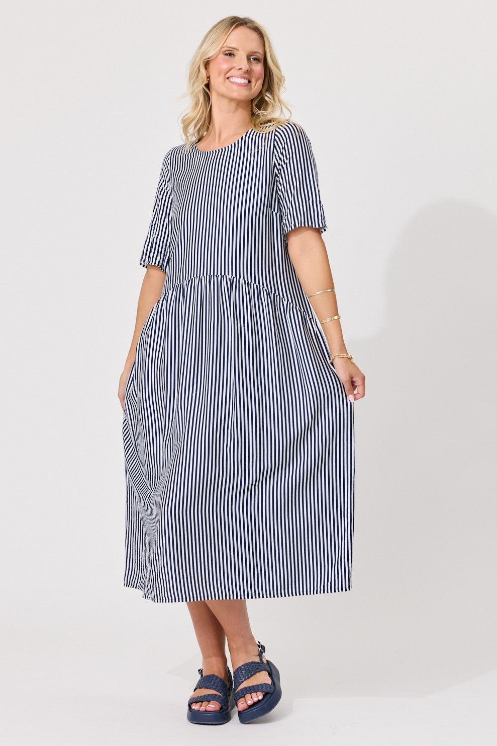 A woman modeling a navy and white vertically striped midi dress with short puff sleeves and a gathered waist, paired with dark blue slide sandals, against a plain light gray studio background.