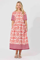 A woman modeling a short-sleeved midi dress with a white base and red-orange floral damask pattern, featuring solid dark pink patterned sleeves and a matching patterned hem, set against a plain light gray studio background.