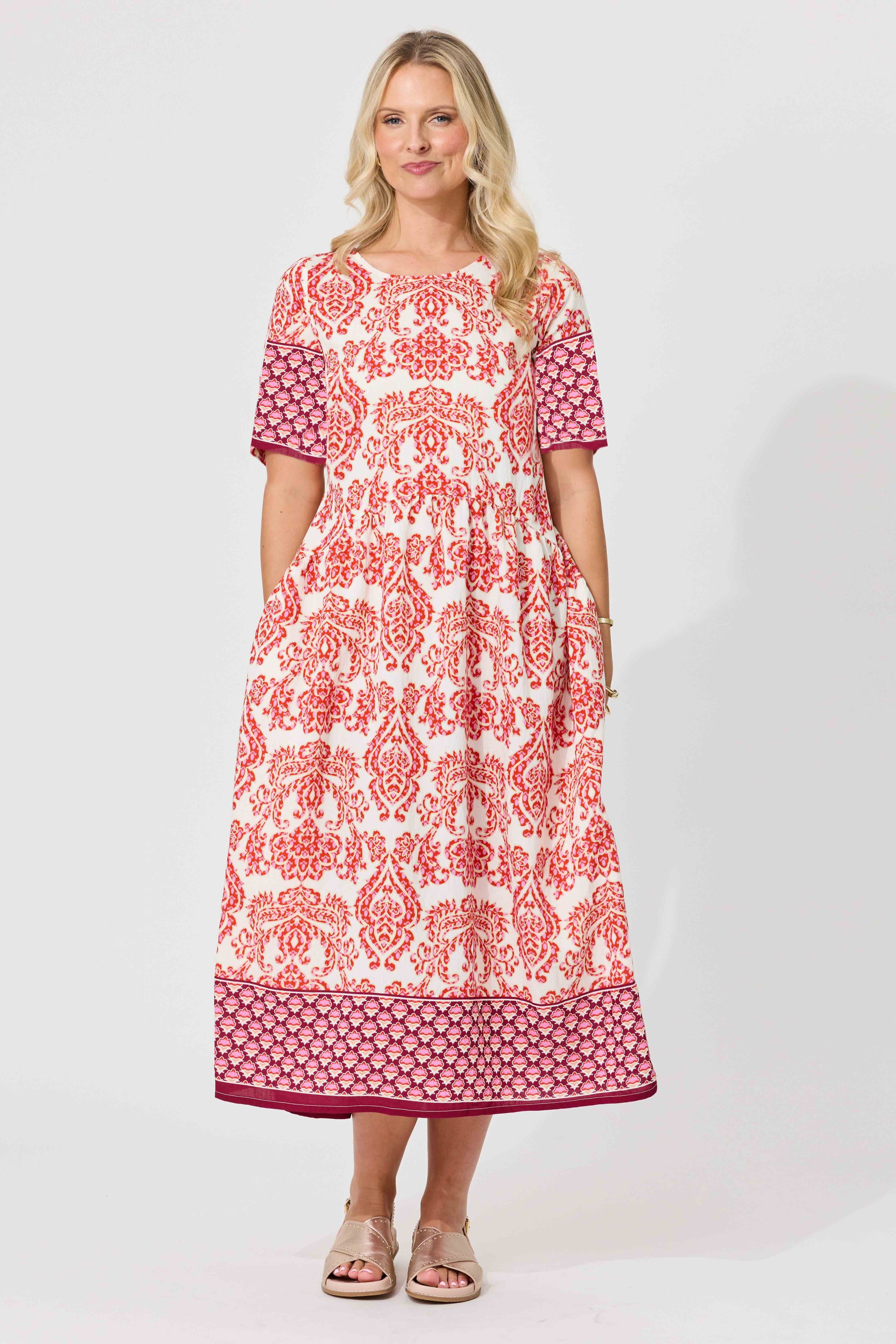 A woman modeling a short-sleeved midi dress with a white base and red-orange floral damask pattern, featuring solid dark pink patterned sleeves and a matching patterned hem, set against a plain light gray studio background.