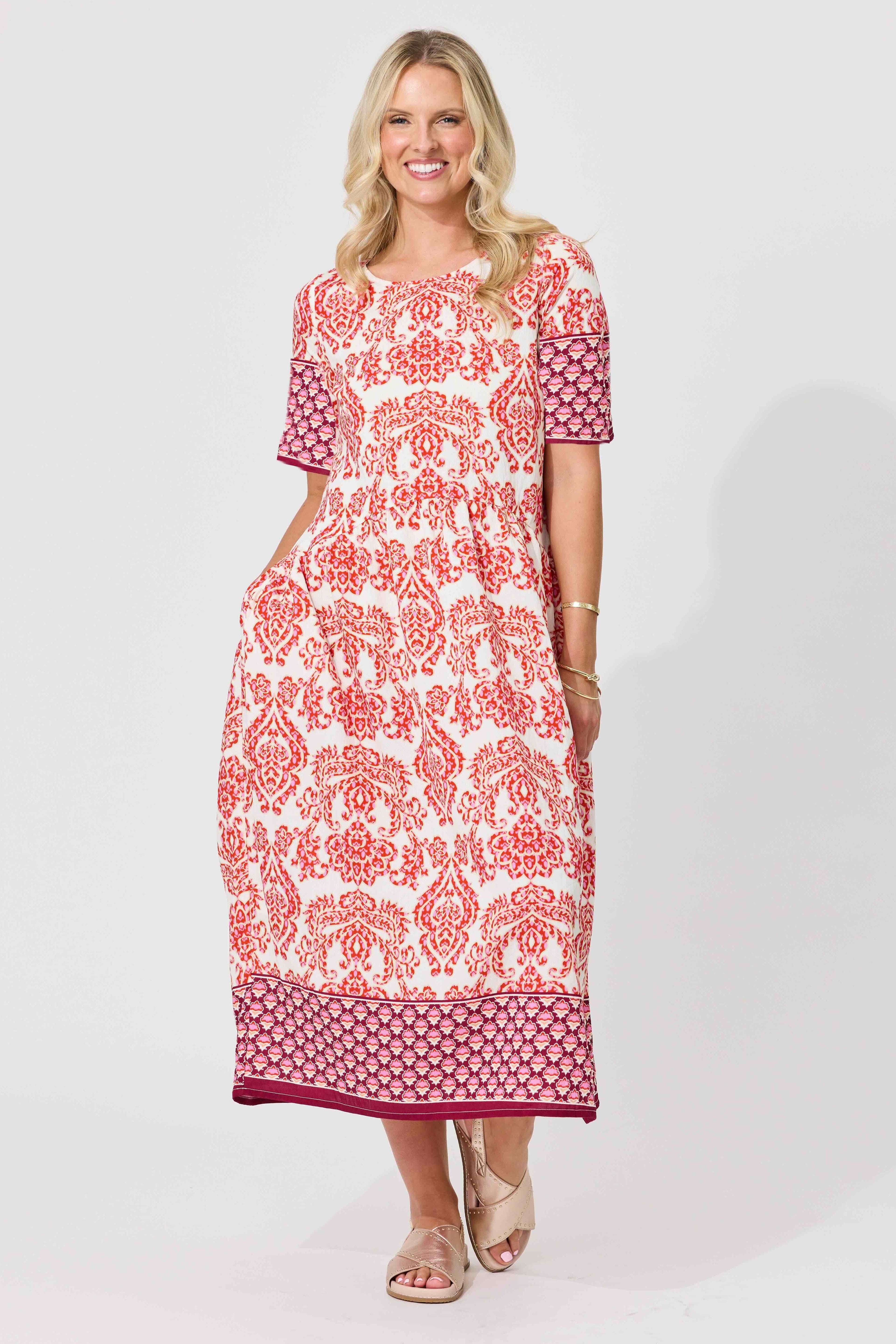 A woman modeling a short-sleeve midi dress with a red and orange floral pattern on a white background, featuring a solid magenta patterned hem and sleeve cuffs, set against a plain light gray studio background.