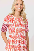 A smiling blonde woman wearing a short-sleeve tunic dress with a red and orange damask pattern on an off-white background, featuring solid red patterned sleeves, set against a plain light background.