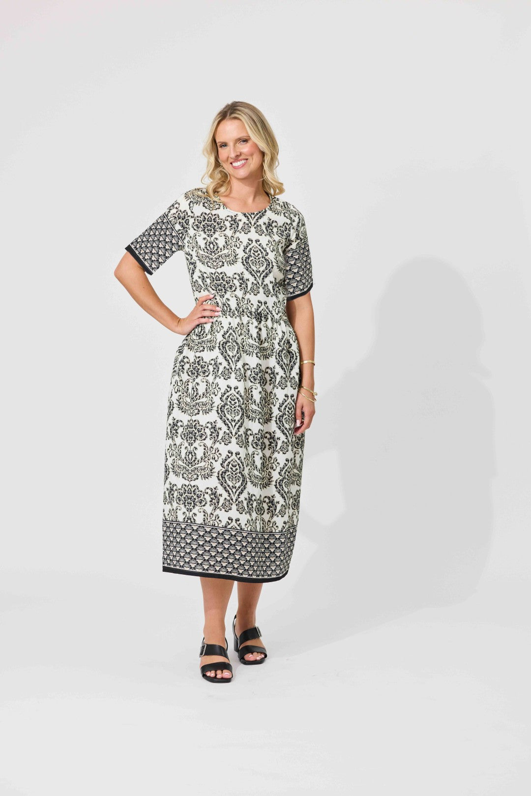 A woman modeling a short-sleeve midi dress with a black and white damask pattern, standing against a light gray studio background.