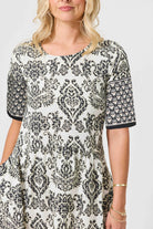 A woman wearing a short-sleeve tunic dress with a black and white damask pattern, featuring solid black cuffs and a scoop neckline, photographed against a light gray studio background.