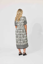 A woman viewed from behind wearing a short-sleeve midi dress with a black and white geometric pattern, paired with black sandals, against a plain light gray studio background.