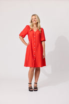 A woman modeling a bright red short-sleeve button-front dress with a V-neck and knee-length skirt, standing against a plain light gray studio background.