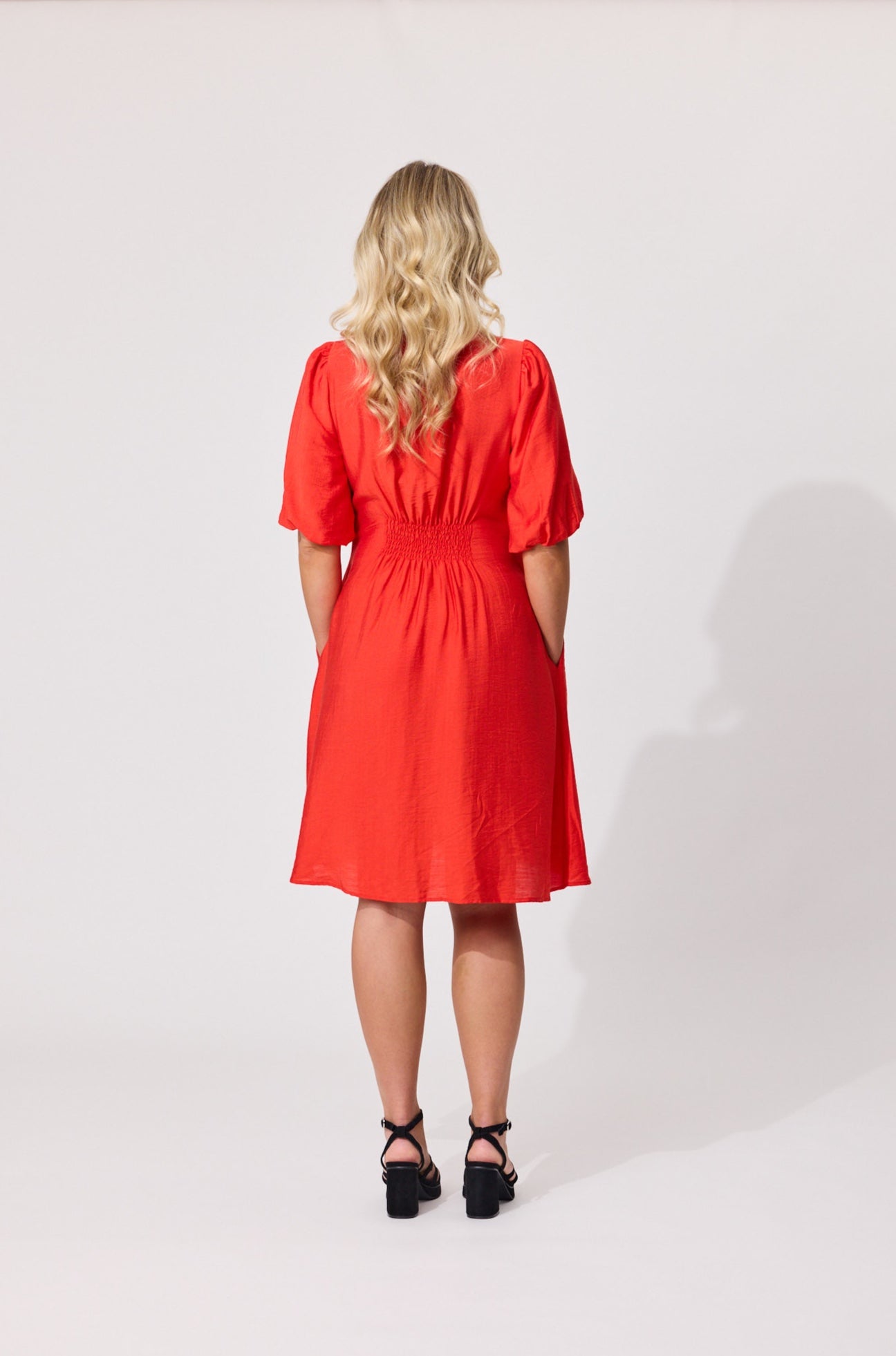 A woman with blonde wavy hair is shown from the back wearing a bright red short-sleeve midi dress with puff sleeves and a gathered waist, paired with black strappy heels, against a light gray studio background.