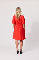 A woman with blonde wavy hair is shown from the back wearing a bright red short-sleeve midi dress with puff sleeves and a gathered waist, paired with black strappy heels, against a light gray studio background.