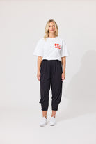 A woman modeling a white short-sleeve t-shirt with a red heart pattern on the left chest, paired with black jogger-style pants and white sneakers, against a plain light gray studio background.
