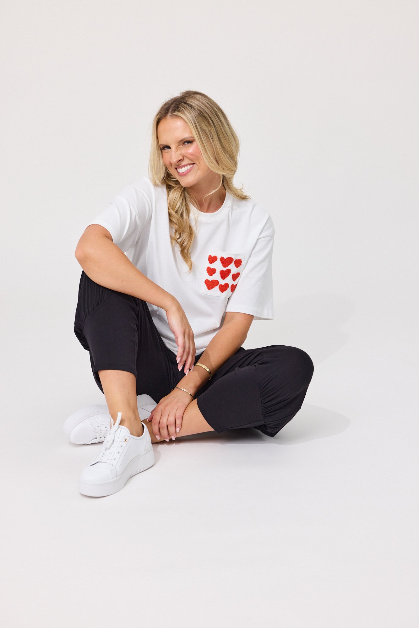 A woman sitting on a light gray studio floor wearing a white t-shirt with a red heart pattern on the chest, black pants, and white sneakers.