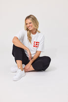 A woman sitting on a light gray studio floor wearing a white t-shirt with a red heart pattern on the chest, black pants, and white sneakers.
