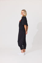 A woman modeling a black two-piece outfit consisting of a short-sleeve cropped top with a red graphic on the chest and matching loose-fitting jogger pants, paired with white sneakers, against a plain white studio background.