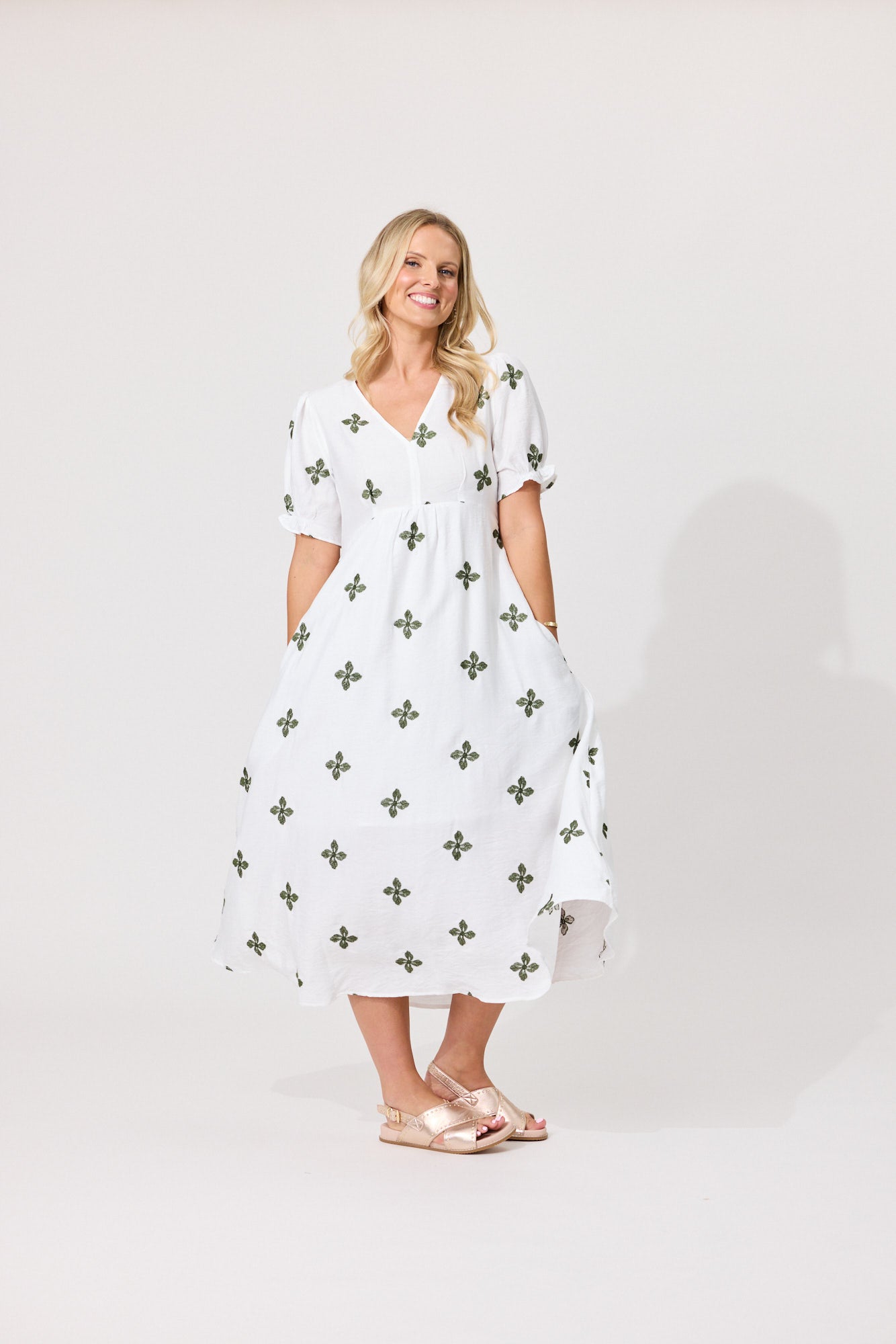 A woman modeling a white midi dress with a green cross pattern, standing against a light gray studio background.