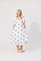 A woman modeling a white midi dress with a green cross pattern, standing against a light gray studio background.