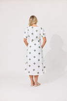 A woman viewed from behind wearing a white midi dress with short puff sleeves and an allover green clover pattern, standing against a plain light gray studio background.