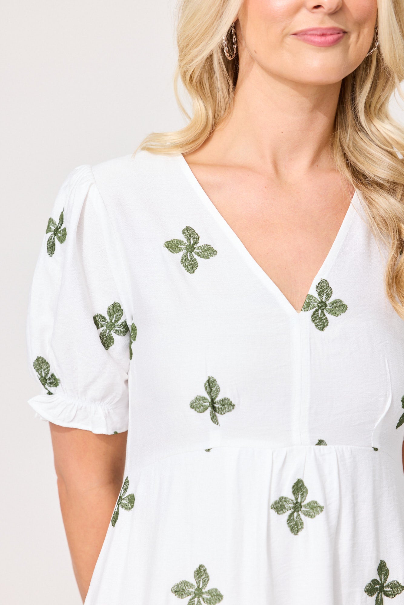 A close-up of a woman wearing a white V-neck dress with short puff sleeves and green embroidered clover motifs scattered across the fabric.