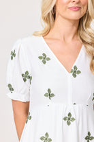 A close-up of a woman wearing a white V-neck dress with short puff sleeves and green embroidered clover motifs scattered across the fabric.