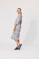A woman modeling a knee-length wrap dress with a gray and white floral pattern, paired with navy blue slide sandals, standing against a plain light gray studio background.