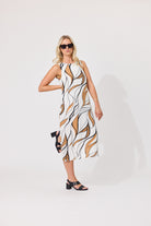 A woman modeling a sleeveless midi dress with an abstract wavy pattern in white, black, and tan, styled with black strappy heels and sunglasses against a plain white studio background.