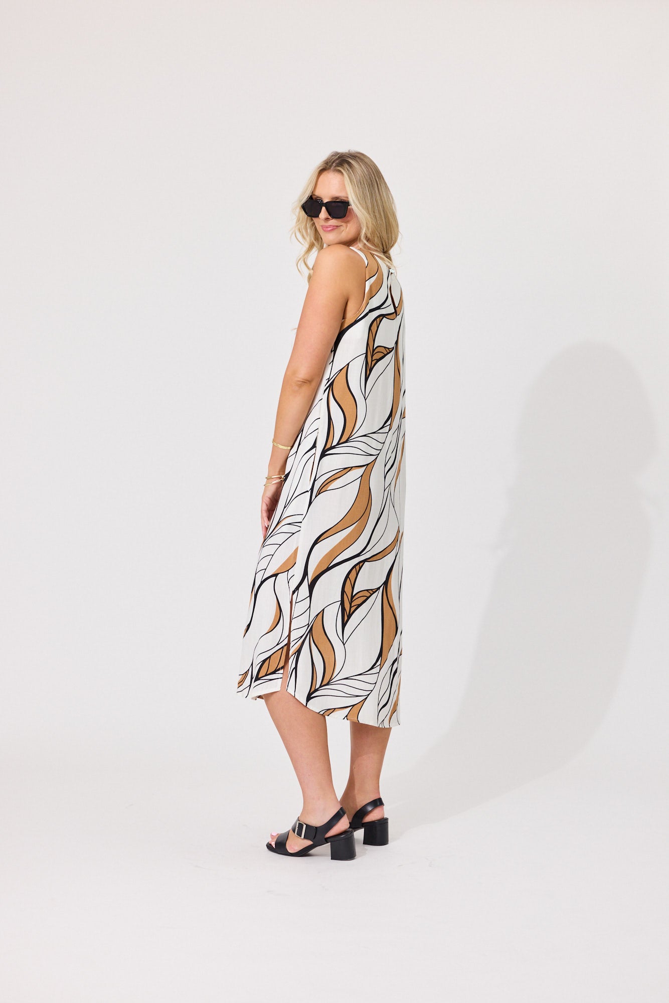 A woman modeling a sleeveless midi dress with a white base and an abstract brown and black leaf-like pattern, photographed against a plain light gray studio background.