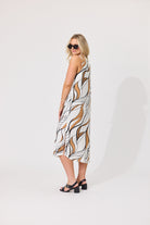 A woman modeling a sleeveless midi dress with a white base and an abstract brown and black leaf-like pattern, photographed against a plain light gray studio background.