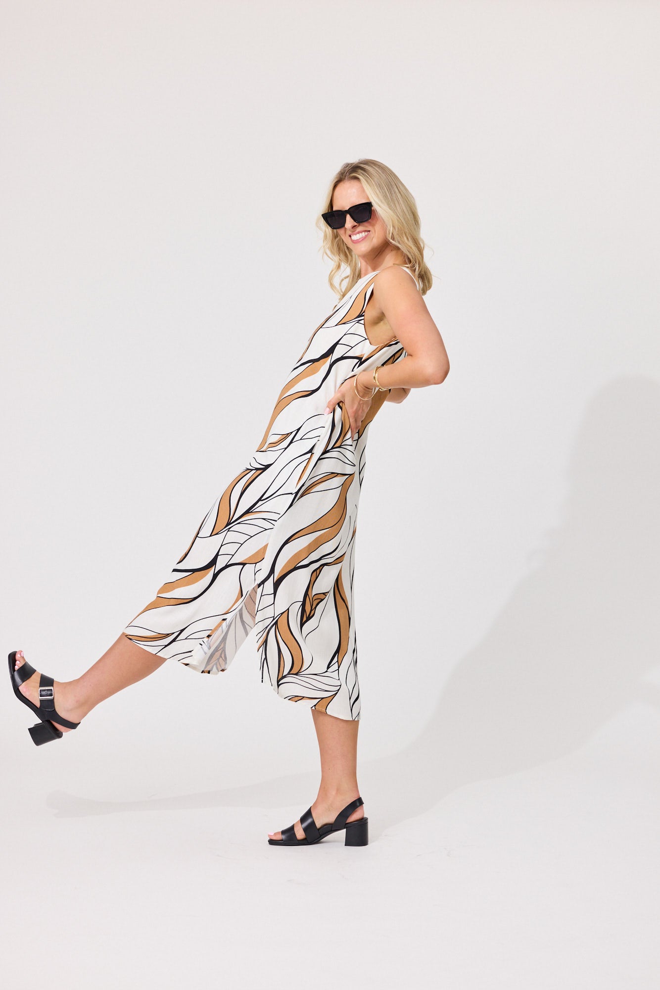 A woman modeling a sleeveless midi dress with a white base and an abstract brown and black line pattern, styled with black sandals and sunglasses against a plain white studio background.