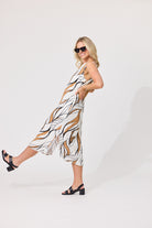 A woman modeling a sleeveless midi dress with a white base and an abstract brown and black line pattern, styled with black sandals and sunglasses against a plain white studio background.