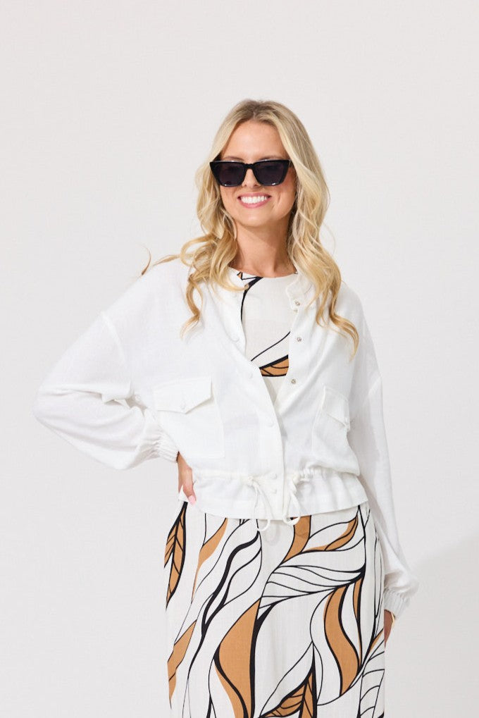 A woman modeling a white long-sleeve button-up shirt with a drawstring waist, paired with a white skirt featuring a brown and black leaf pattern, set against a plain white studio background.