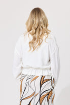 A woman viewed from behind wearing a white long-sleeve top with a ruffled waist and a white skirt featuring a brown and black leaf pattern, set against a plain light gray background.