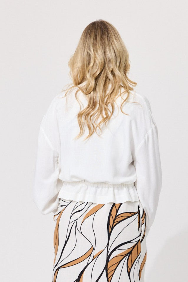 A woman viewed from behind wearing a white long-sleeve top with a ruffled waist and a white skirt featuring a brown and black leaf pattern, set against a plain light gray background.
