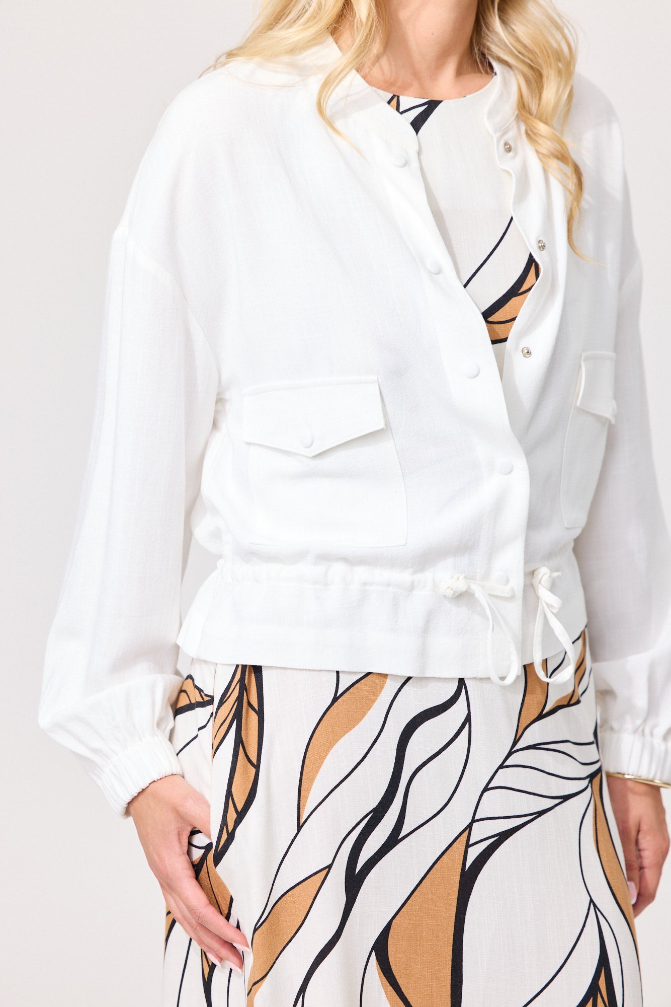A woman wearing a white long-sleeve button-front jacket with flap chest pockets and a drawstring waist, paired with a white skirt featuring a tan and black abstract leaf pattern, against a light studio background.