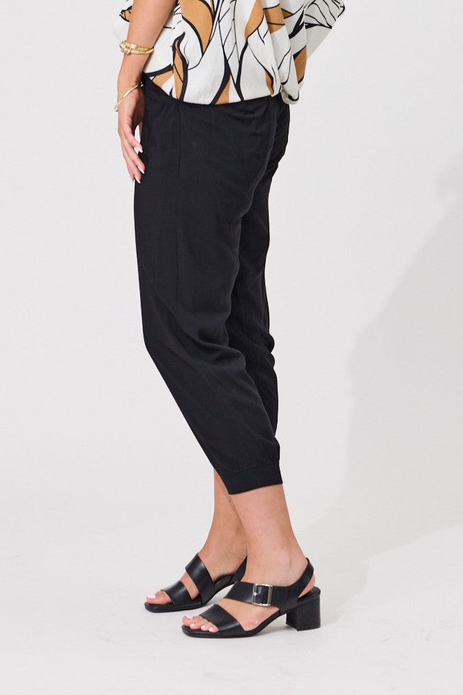 A model wearing black cropped trousers with a relaxed, tapered fit and side pockets, paired with black block-strap sandals and a patterned top, shown from the waist down against a white studio background.