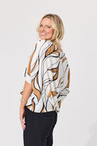 A woman wearing a white short-sleeve blouse with a brown and black abstract leaf pattern, paired with black pants, photographed against a light gray studio background.