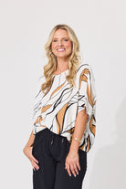 A woman modeling a white short-sleeve blouse with a brown and black abstract wavy pattern, paired with black pants, against a plain light gray background.