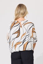A woman viewed from the back wearing a white blouse with a brown and black abstract leaf-like pattern and black pants, set against a plain light gray studio background.
