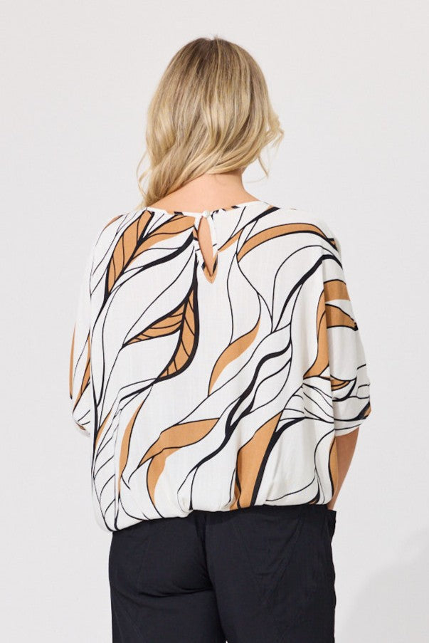 A woman viewed from the back wearing a white blouse with a brown and black abstract leaf-like pattern and black pants, set against a plain light gray studio background.