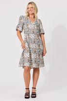 A woman modeling a short-sleeve V-neck dress with a multicolor geometric patchwork print, paired with black strappy sandals, against a plain light gray studio background.