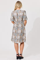 A woman viewed from the back wearing a knee-length dress with puff sleeves and an allover patchwork-style print in beige, white, blue, and brown tones. She is standing against a plain light gray studio background and wearing black high-heeled sandals.