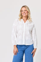 A woman wearing a white long-sleeve blouse with delicate floral embroidery and a drawstring neckline, paired with blue jeans, standing against a light gray studio background.