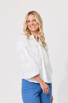 A smiling blonde woman wearing a white embroidered blouse with three-quarter sleeves and blue jeans, standing against a plain light gray studio background.