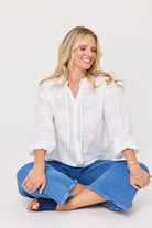 A smiling blonde woman sits cross-legged on a light gray studio background, wearing a white embroidered long-sleeve blouse with drawstrings and blue cropped jeans.