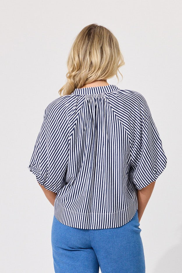 A woman viewed from the back wearing a navy and white vertically striped blouse with puff sleeves and blue pants, against a plain light gray studio background.